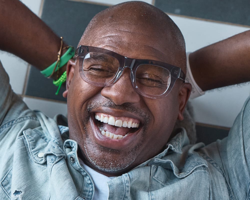 A close-up shot of a smiling man wearing clear-framed eyeglasses and a light blue denim shirt, with his arms behind his head against a tiled wall.