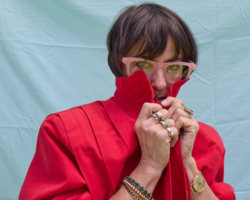 A person wearing a bright red high-neck jacket or dress, pink cat-eye sunglasses, and multiple gold rings, holding the collar up around their face against a light blue backdrop.