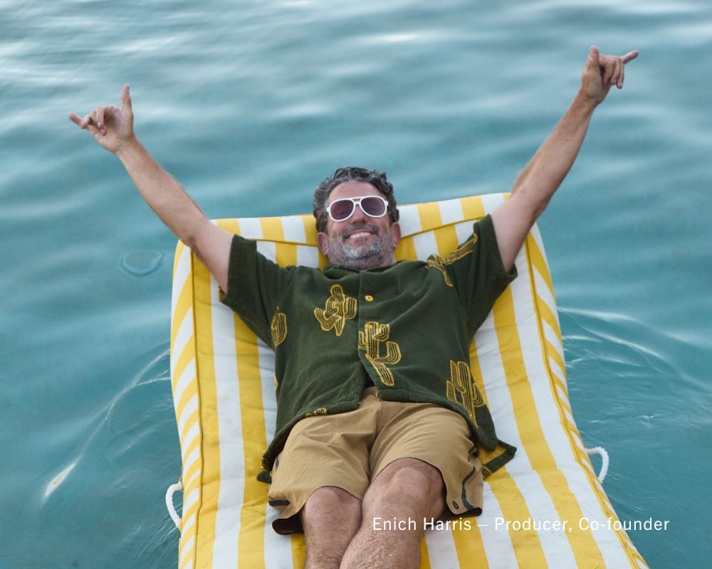 A man wearing sunglasses and a green cactus-print short-sleeve shirt lies on a yellow and white striped lounge chair on blue water, with his arms raised in the air.